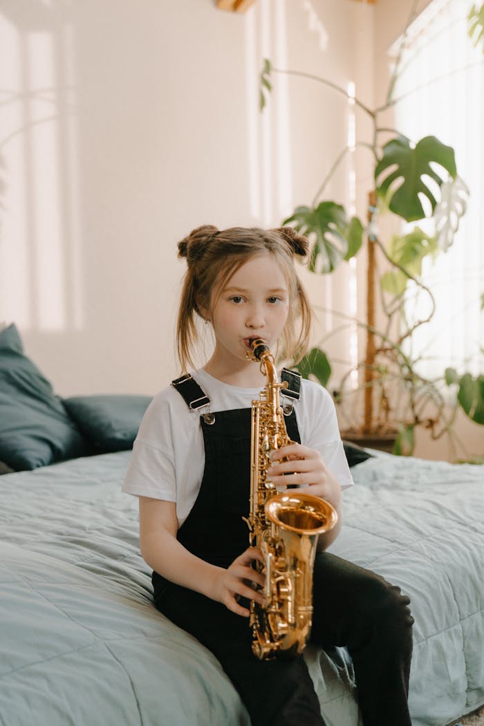 A young girl sits on a bed playing a saxophone, surrounded by a warm, cozy interior with plants.